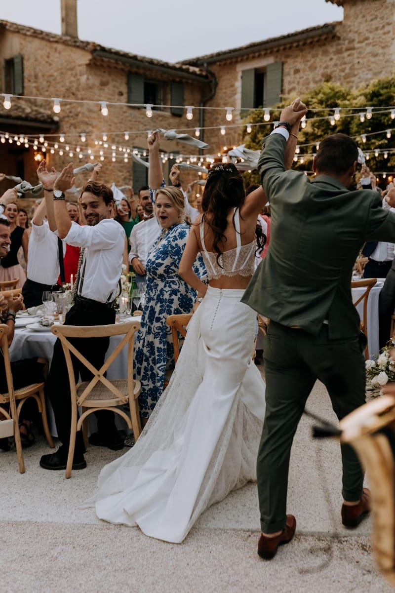 Bride and groom enter outdoor reception at Provencal farmhouse courtyard as guests cheer under Edison bulb lights