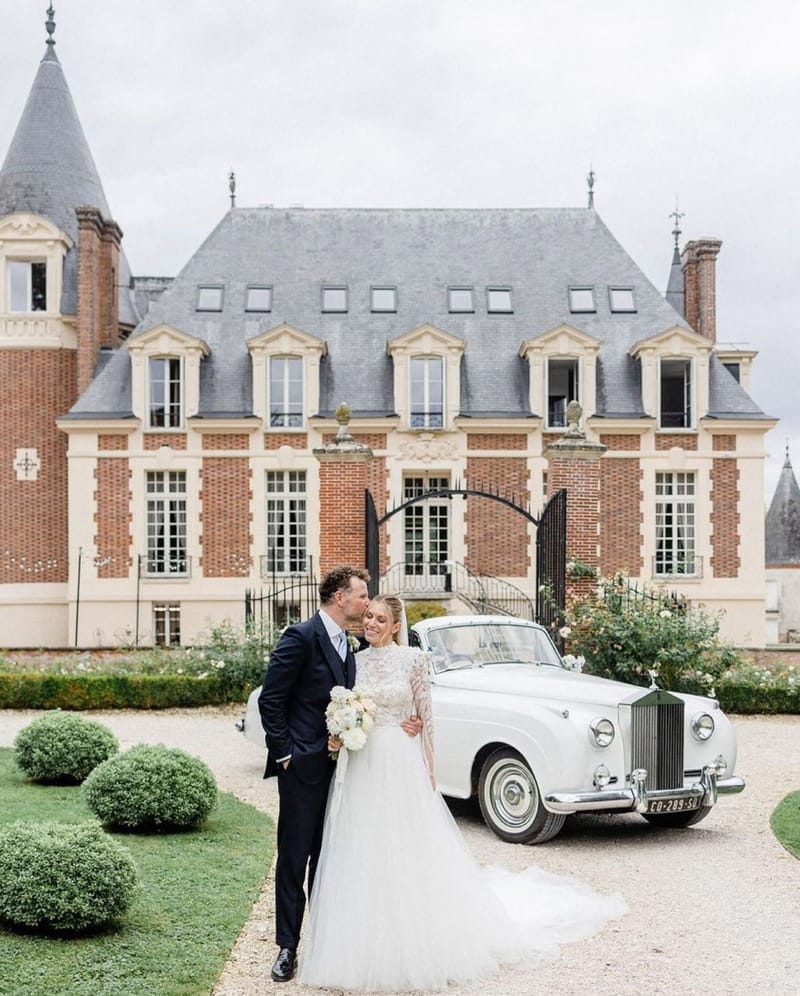 Groom kisses bride on cheek in front of red brick French chateau with vintage white Rolls-Royce