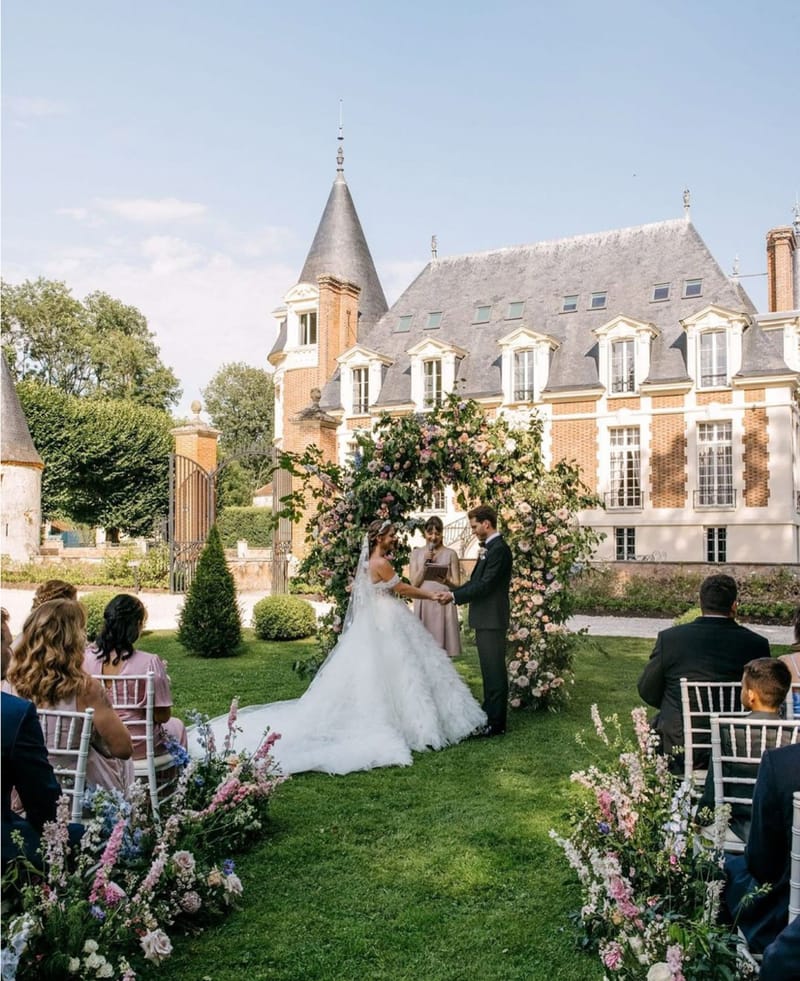 Outdoor chateau ceremony with circular floral arch of pink roses, guests seated in white chairs