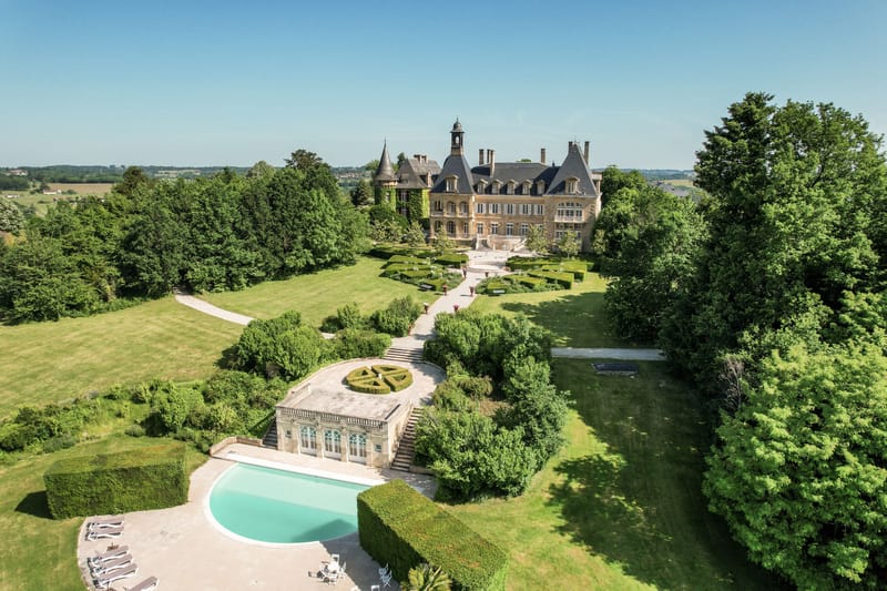 Aerial golden limestone chateau with bell tower, curved pool, box parterre, and orangery pool house