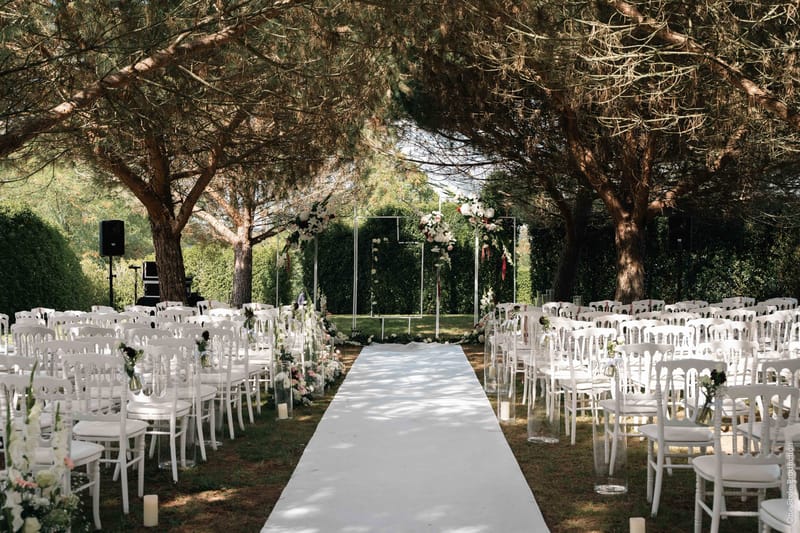 Ceremony setup with square metal arch, white and blush florals, and Napoleon chairs under pines