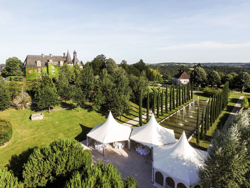 Aerial ivy-covered chateau with slate turrets, white marquee tents, fountain canal, and cypress rows on estate grounds