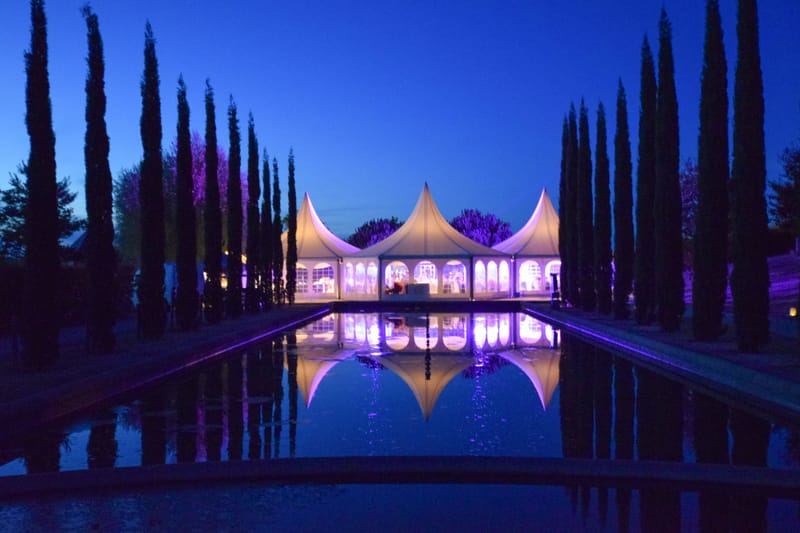 White marquee tent lit from within at dusk reflected in ornamental pool flanked by cypress trees with purple uplighting