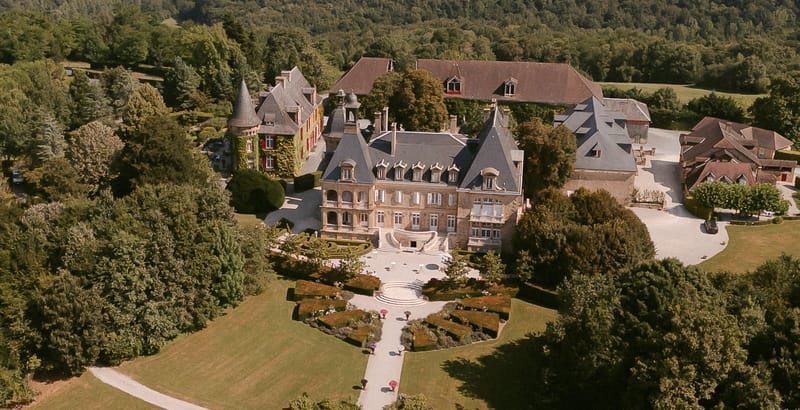 Aerial drone view of French chateau estate with slate mansard roofs, formal geometric gardens, and woodland surroundings