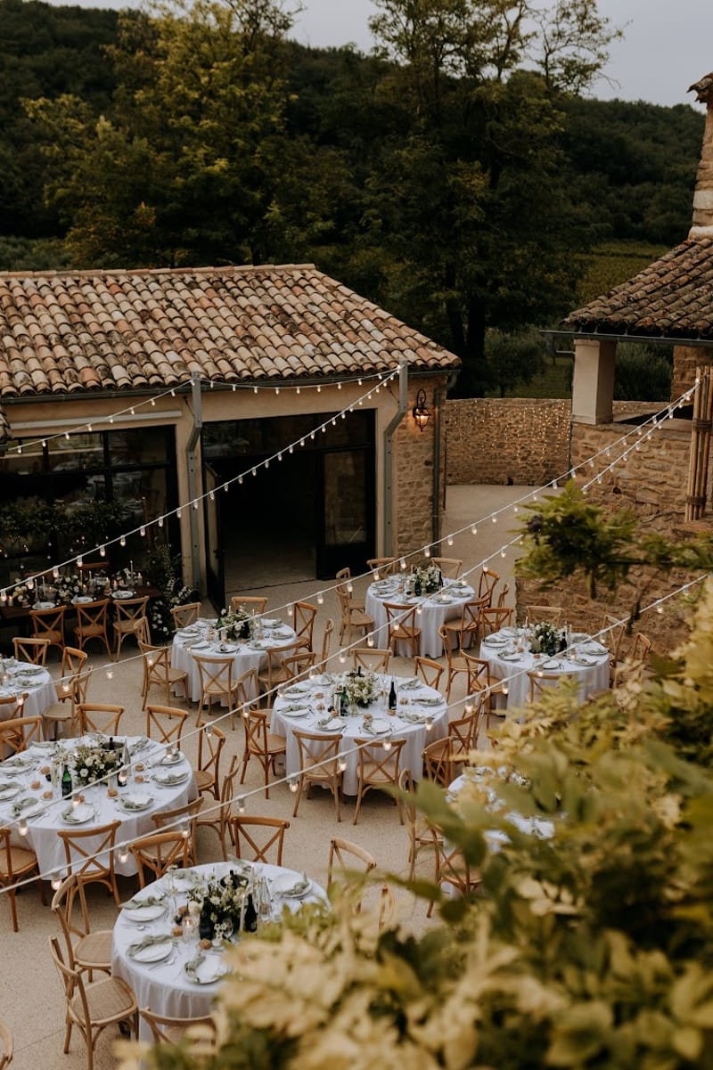 Eight round tables with white linen cross-back chairs and white floral centerpieces under bistro lights in Provencal court...