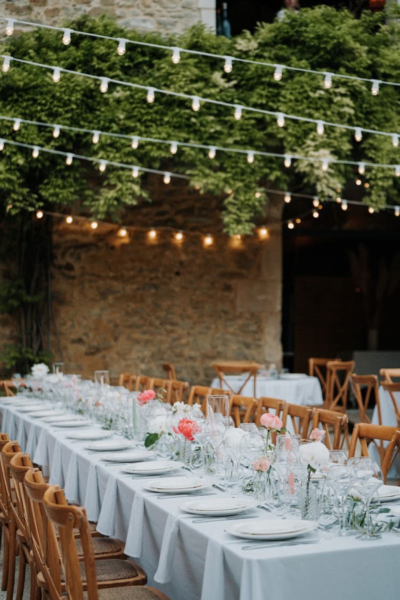 Long reception table with blue-grey linen, coral peonies, and string lights in vine-covered stone courtyard