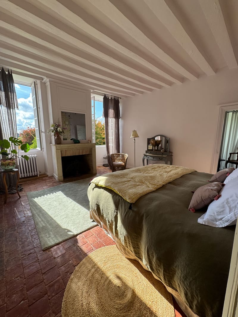French country bedroom with exposed beams, stone fireplace, olive linen bed, and rattan dressing table