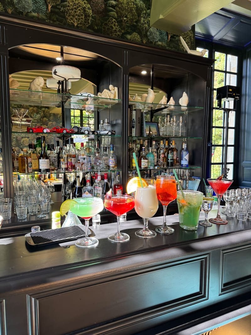 Six colorful cocktails on dark bar counter before mirrored cabinet with spirits and whimsical figurines
