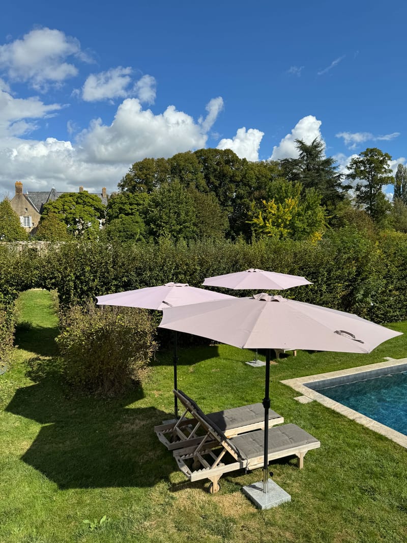 Poolside with dusty pink parasols and wooden loungers before stone manor with slate rooflines and hedging