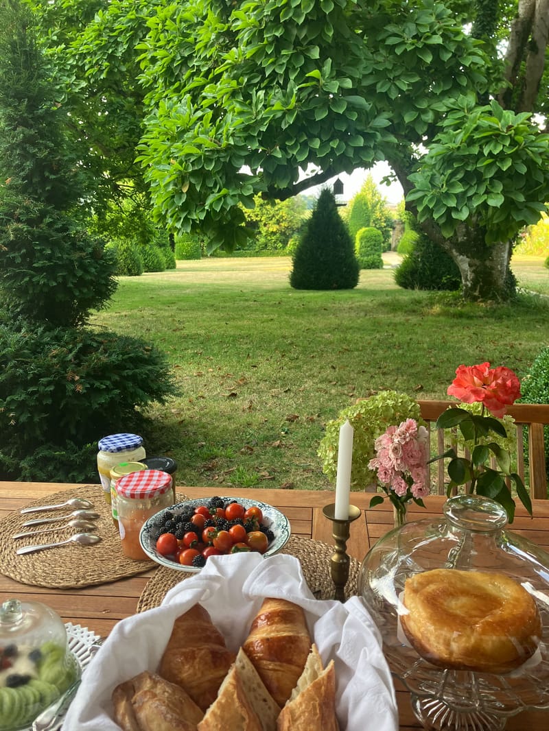 French breakfast spread with croissants, jam, berries, and pink roses on a wooden terrace overlooking a garden