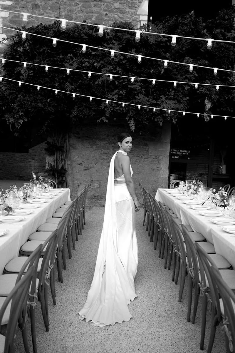 Black-and-white portrait of bride in halter gown standing between long reception tables in stone courtyard