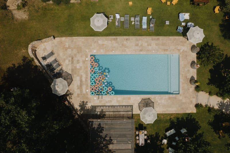 Aerial rectangular pool with colorful inflatable rings, market umbrellas, and grey loungers on surrounding lawn
