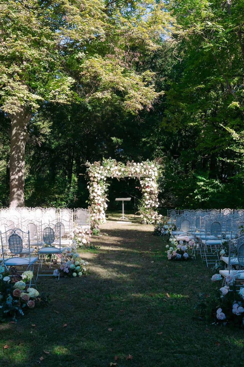 Outdoor garden ceremony setup with floral arch and wrought-iron chairs under mature trees