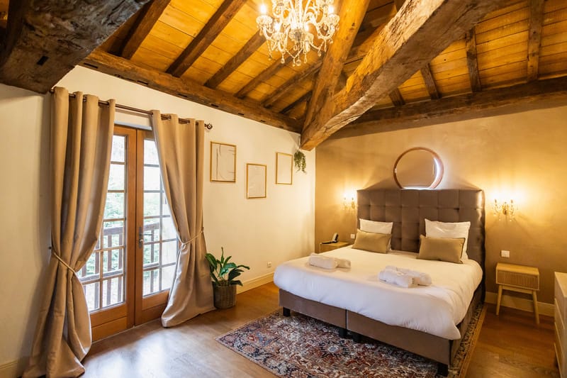 Guest bedroom with exposed beams, crystal chandelier, tufted grey headboard, and French doors to balcony