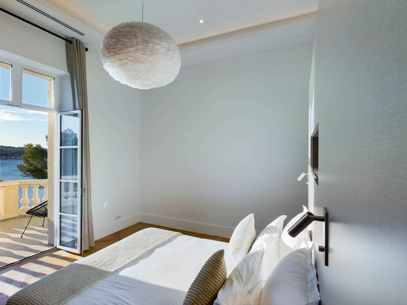 Guest bedroom with white linen bed, feather pendant light, and French doors opening to balcony with water view