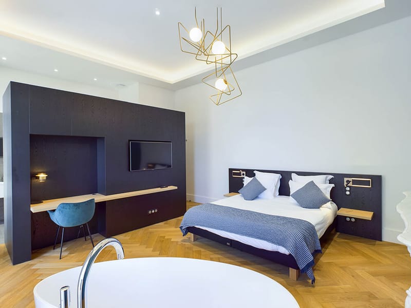 Hotel guest suite with freestanding oval bathtub, navy headboard, herringbone oak floor, and gold pendant light
