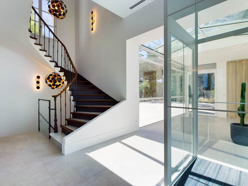 Contemporary venue entrance hall with curved walnut staircase, gold pendant lights, and floor-to-ceiling glass partition