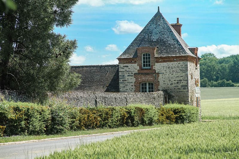 French country estate gatehouse with pointed slate roof, stone and red brick facade, and arched teal-framed windows