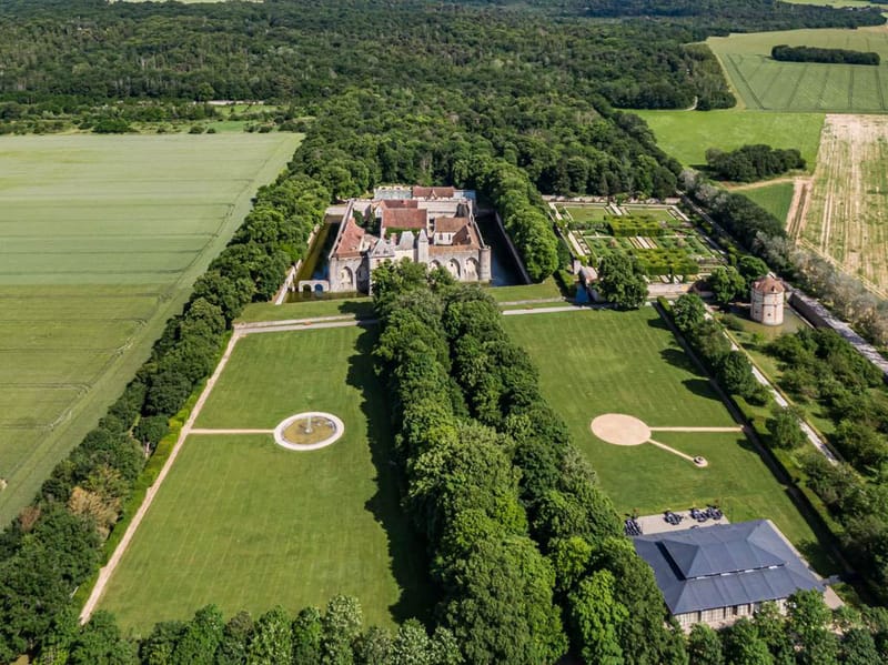 Aerial drone view of French chateau estate with moat, formal gardens, manicured lawns, and reception barn surrounded by wo...