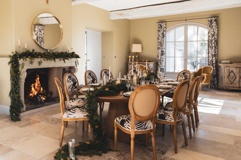 Winter dining room with pine and dried citrus garland, active fireplace, navy leaf-print chairs, and gold mirror