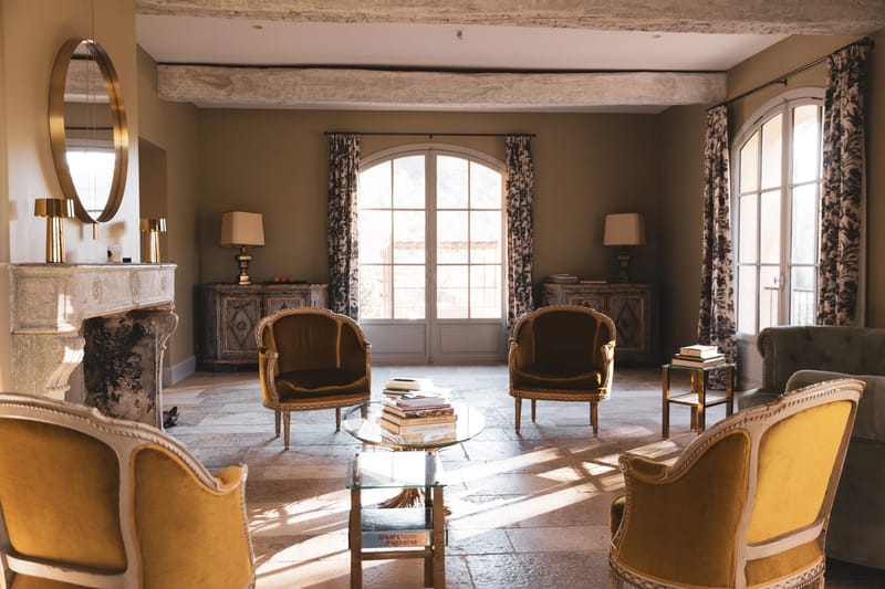 Chateau sitting room with mustard armchairs, stone fireplace, brass mirror, and arched French doors