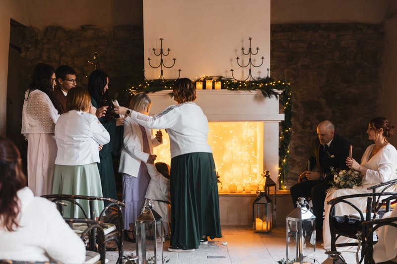 Indoor candlelit ceremony with couple seated as guests pass taper candle before fairy-lit fireplace with greenery
