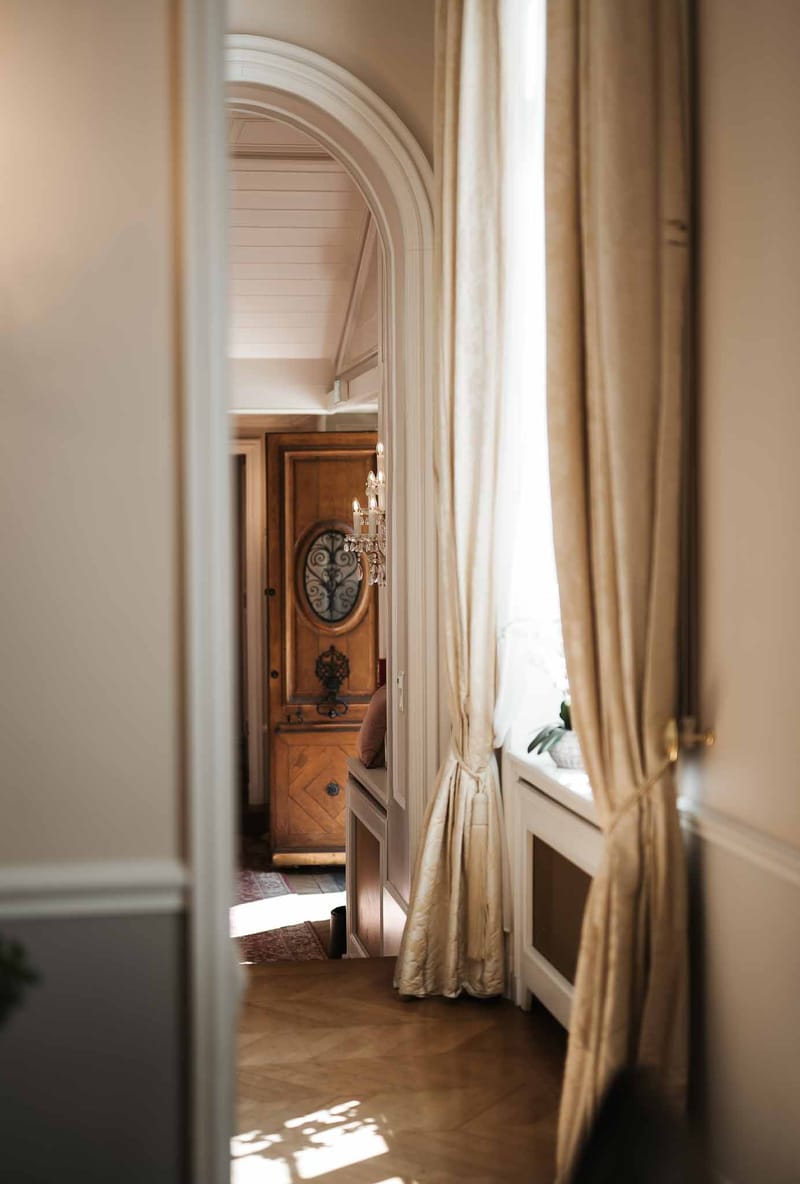Interior view through an arched doorway showing antique armoire, herringbone parquet floor, and silk curtains