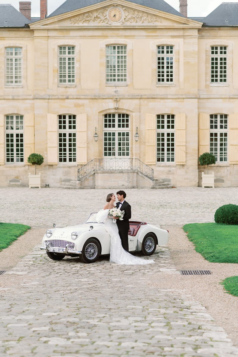 Bride and groom beside vintage white Triumph convertible in front of classical French chateau facade