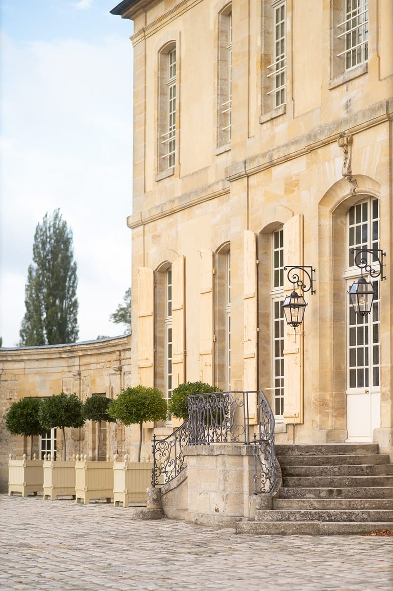 Cream limestone French chateau exterior with arched windows, iron railings, and topiary planters in courtyard