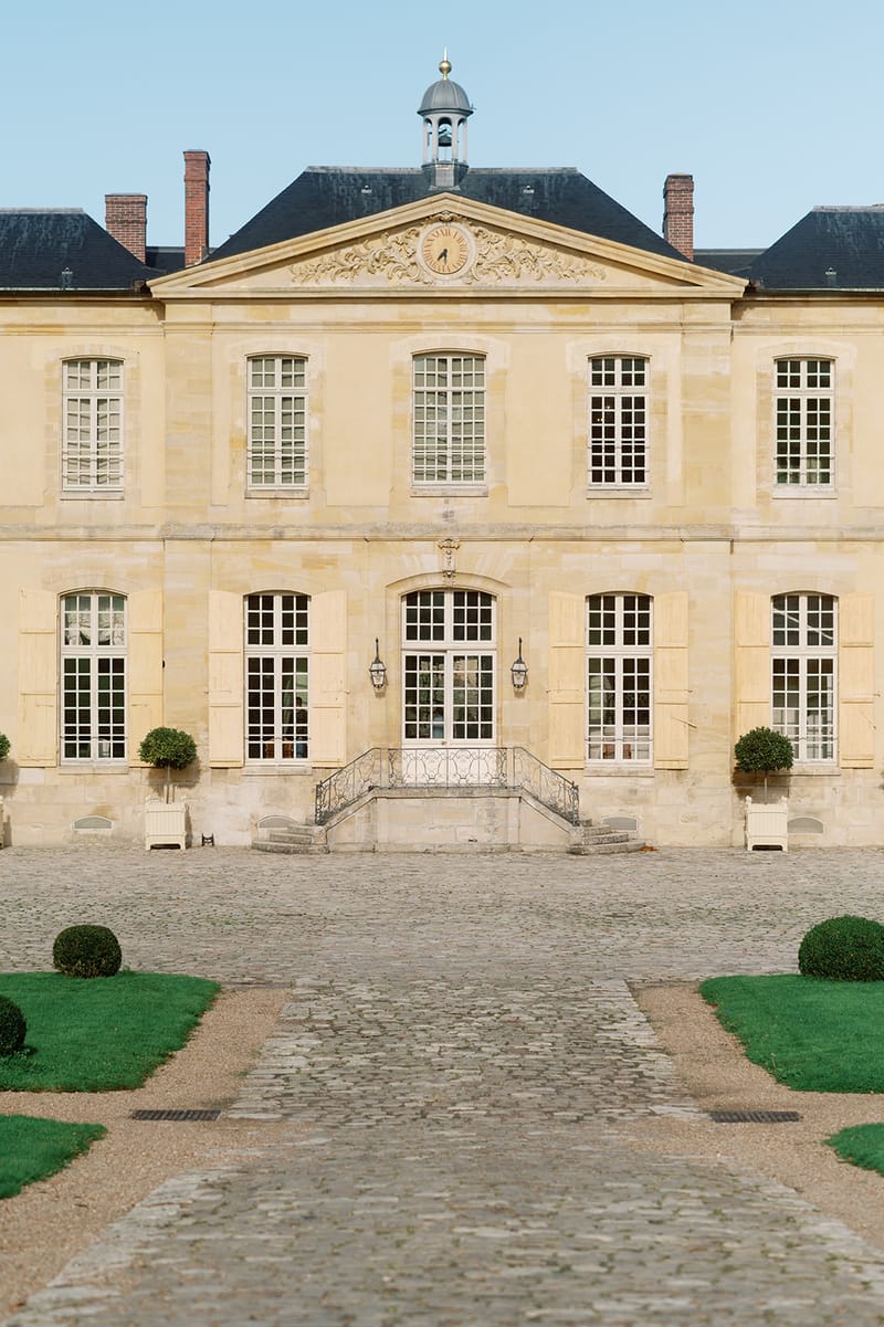 Pale limestone French chateau facade with mansard roof, central clock pediment, and symmetrical box topiaries