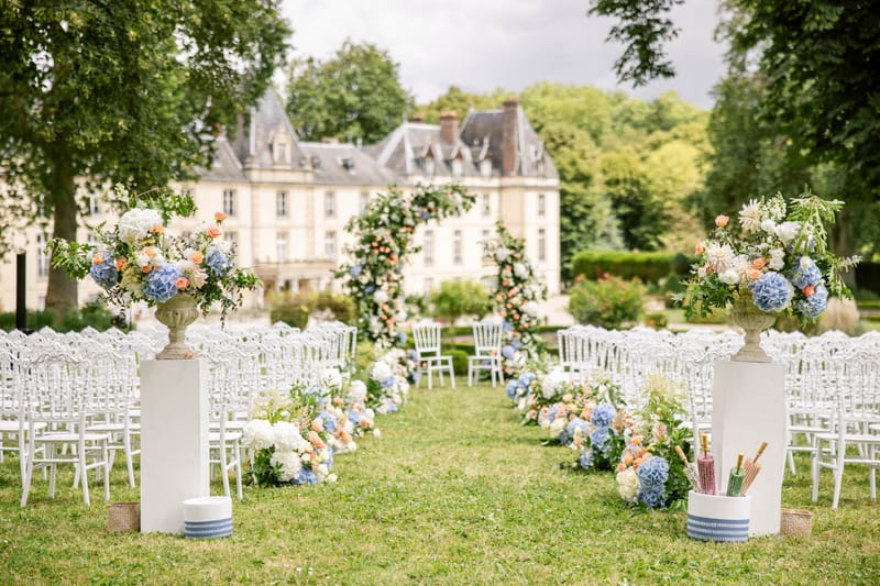 Ceremony aisle with blue hydrangea urns, coral rose ground clusters, and circular greenery arch before chateau