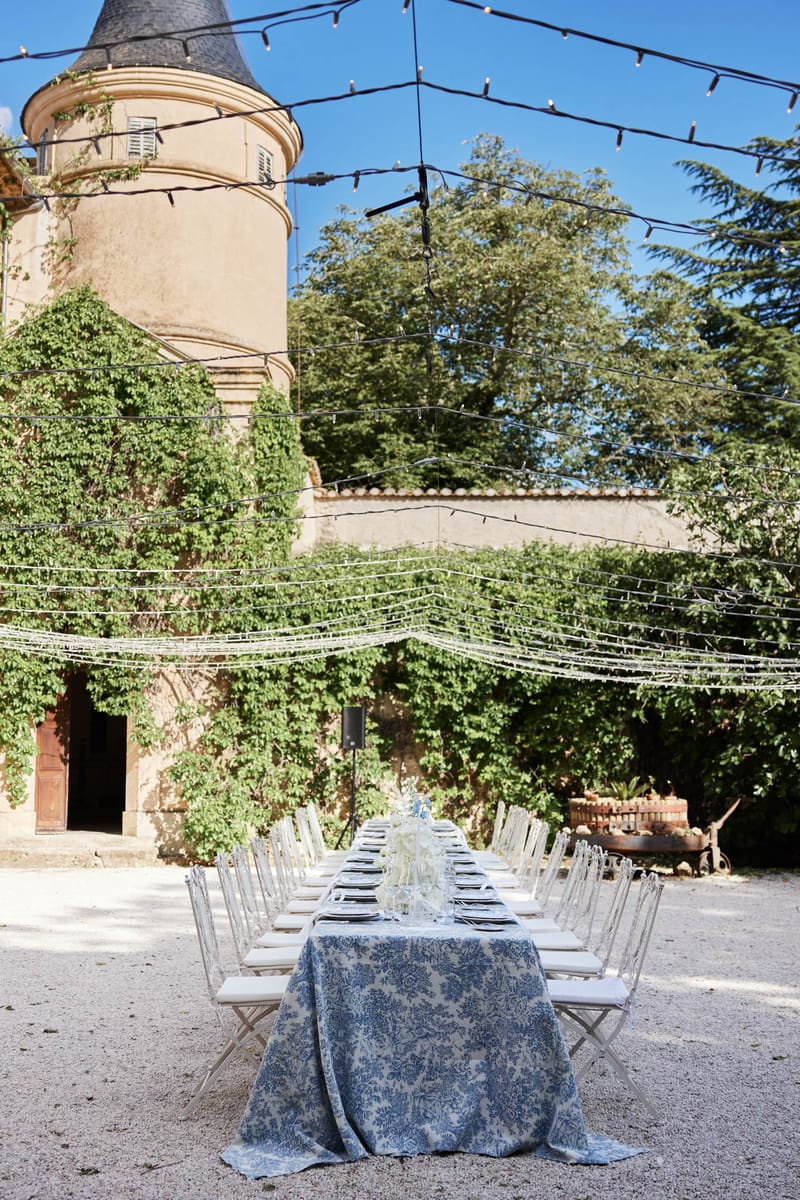 Long reception table with blue damask cloth and ghost chairs in chateau courtyard with fairy lights and stone tower