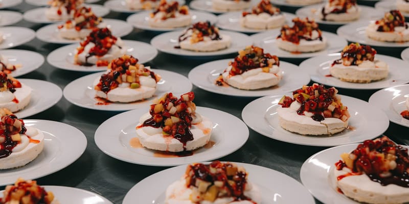 Rows of individual pavlova desserts with mixed berry compote and coulis on stainless steel prep surface
