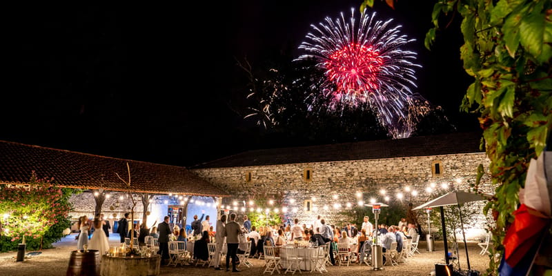 Eighty guests at round tables in stone courtyard with fireworks overhead and Edison lights at night