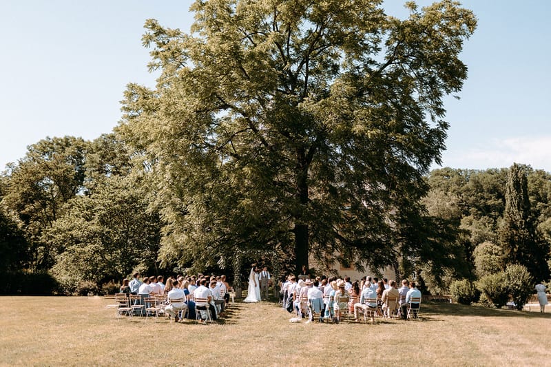 Outdoor wedding ceremony under a large tree with guests seated on wooden chairs on a chateau lawn