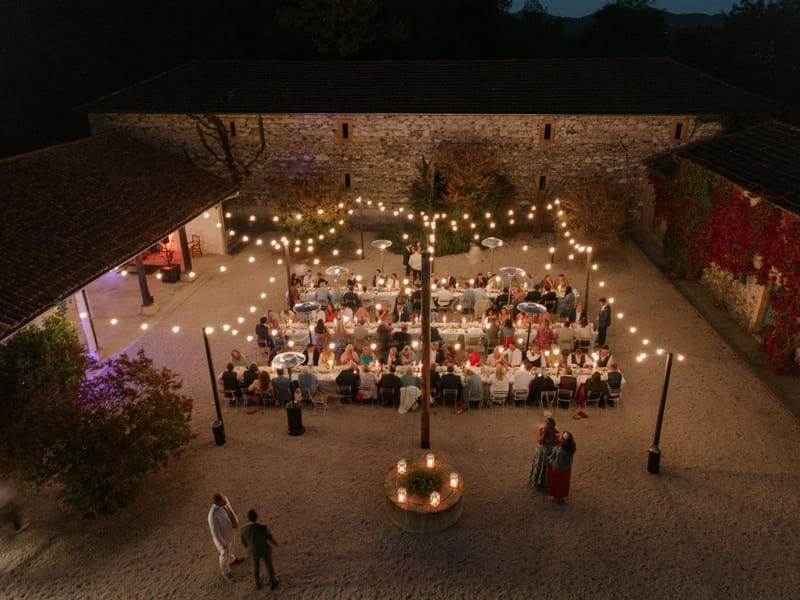 Aerial view of outdoor evening wedding reception in a gravel courtyard with string lights and guests seated at long tables
