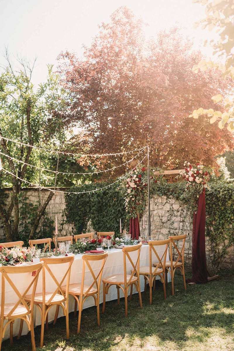 Outdoor reception table with burgundy napkins and blush rose garland under fairy lights in ivy courtyard