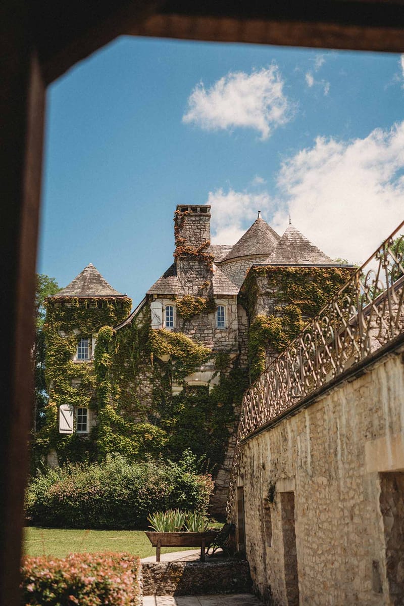 Ivy-covered chateau with conical turrets and wrought-iron staircase framed through doorway on manicured lawn