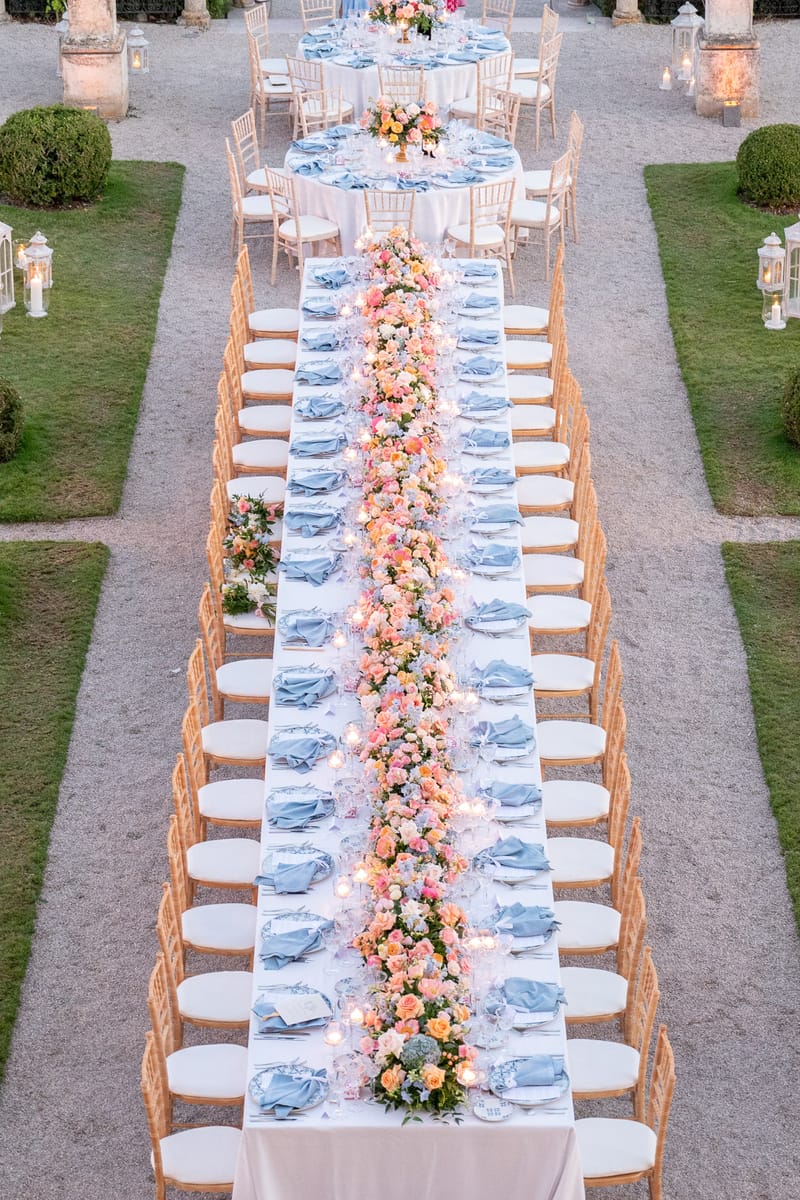 Long banquet table with coral and blush rose runner, dusty blue napkins, and votive candles in formal French garden