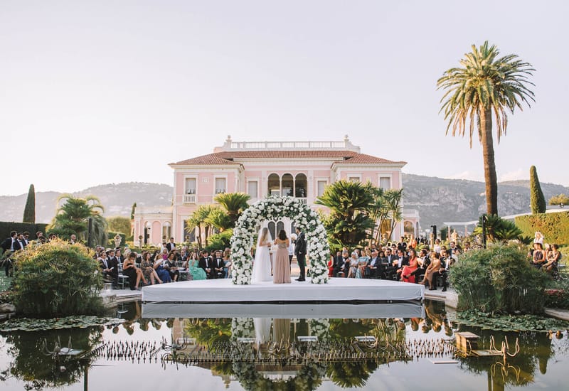 Ceremony on floating platform over reflecting pool with circular white floral arch and pink villa