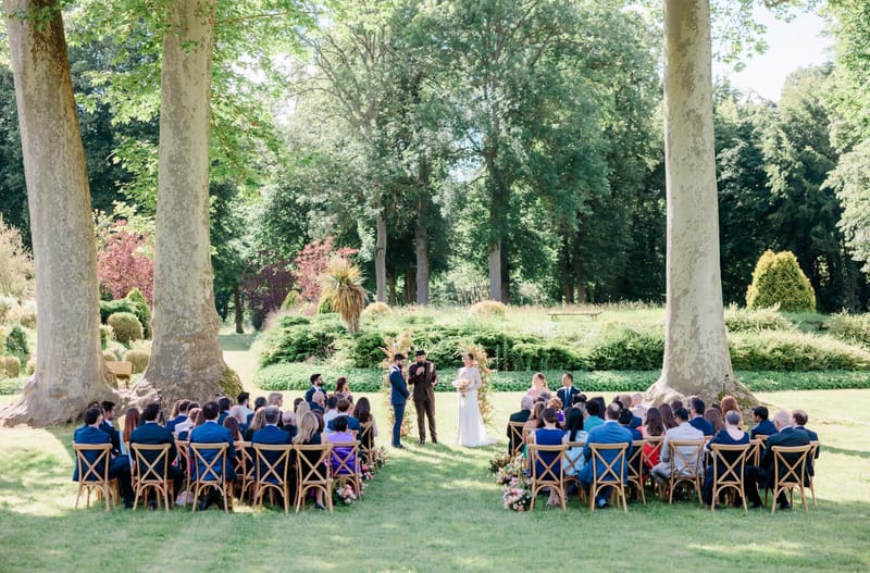 Outdoor wedding ceremony under plane trees with 60 guests, cross-back chairs, and a pink floral arch