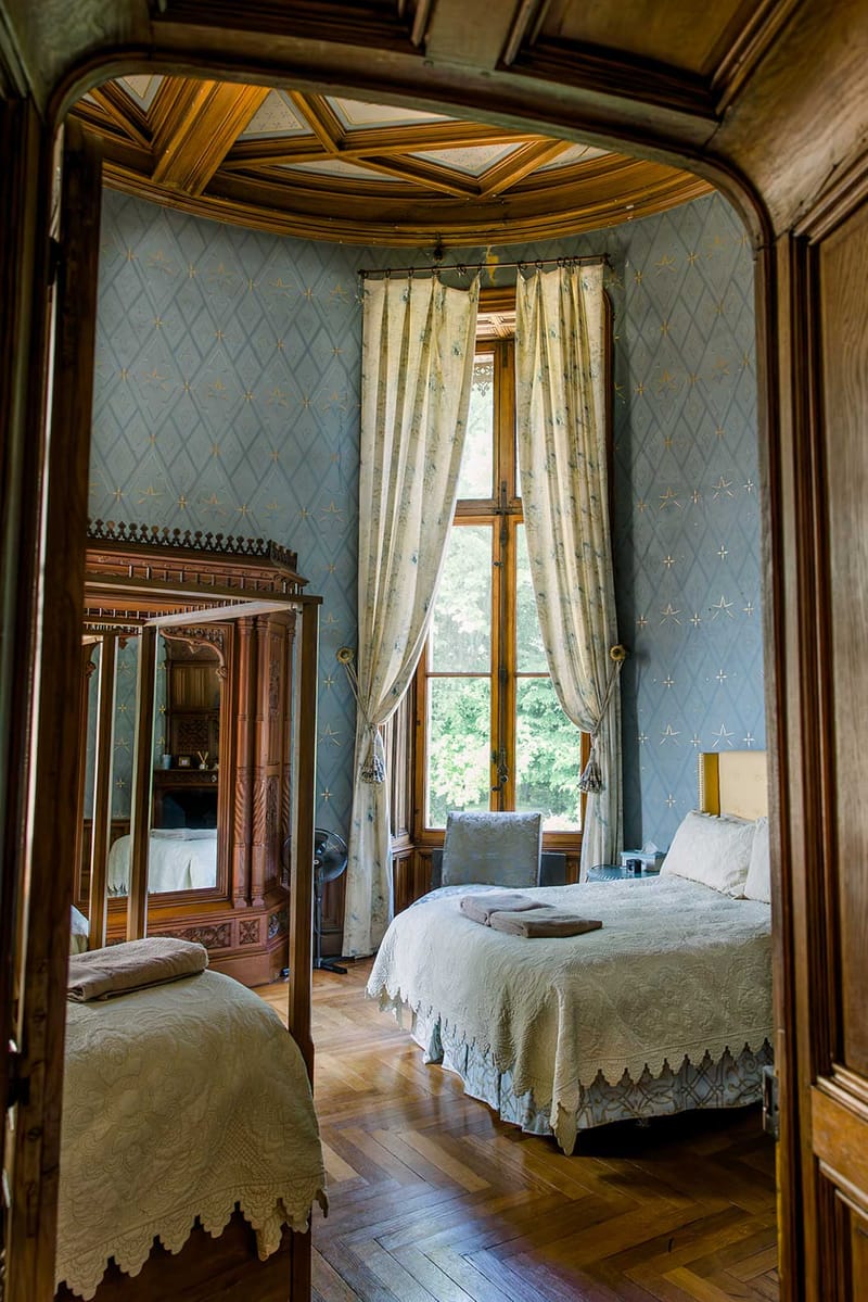 Chateau bedroom with powder blue wallpaper, coffered ceiling, mirrored armoire, and lace-trimmed beds