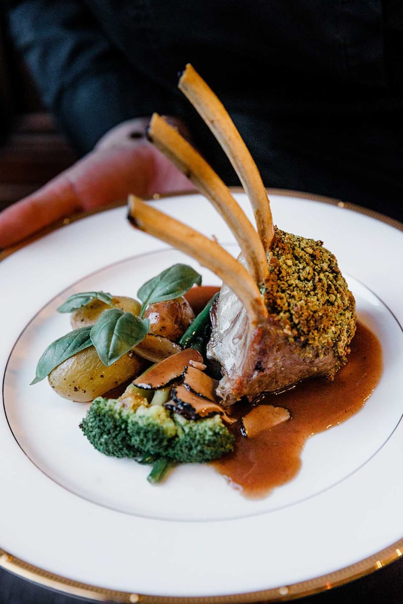 Plated herb-crusted rack of lamb with roasted potatoes and vegetables on gold-rimmed white plate at wedding reception