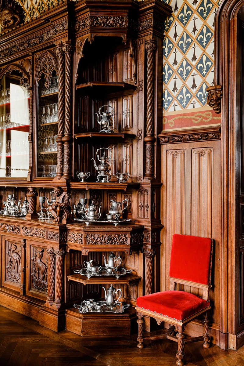 Carved walnut buffet with silver tea sets in a chateau room with herringbone parquet and fleur-de-lis frieze