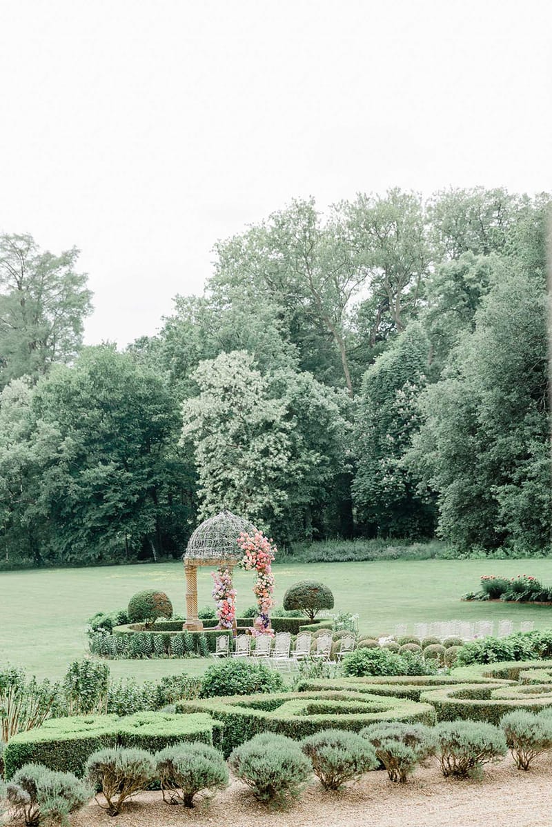 Formal garden ceremony with wire gazebo coral pink and fuchsia floral arch white chairs and boxwood hedges