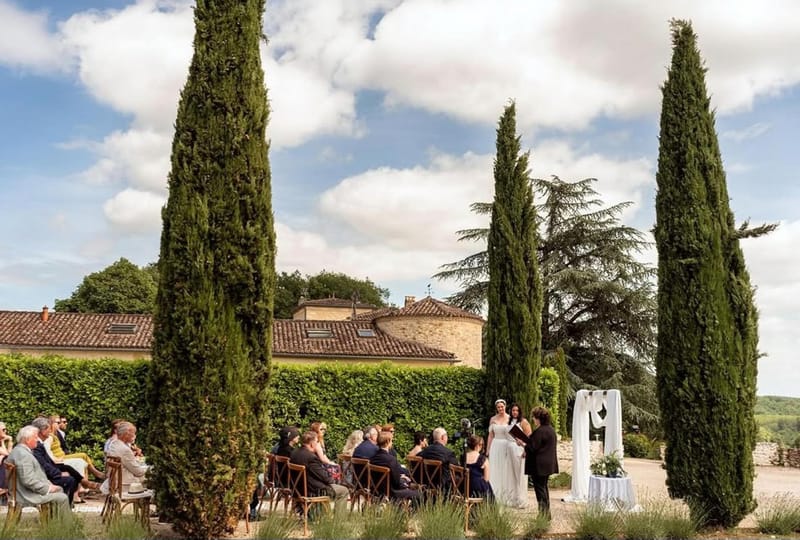 Outdoor wedding ceremony at French chateau with guests seated on wooden chairs facing couple at white-draped altar