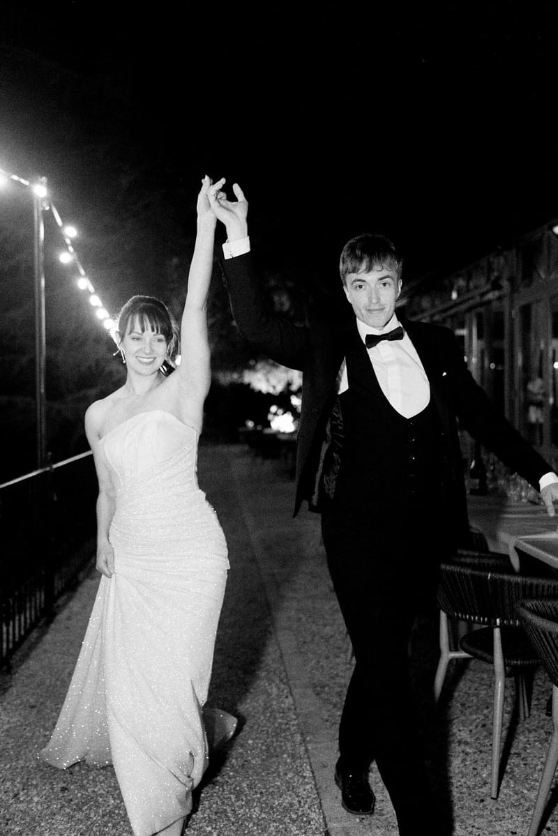 Black and white of couple walking with raised hands under string lights at night reception in fitted gown and tuxedo