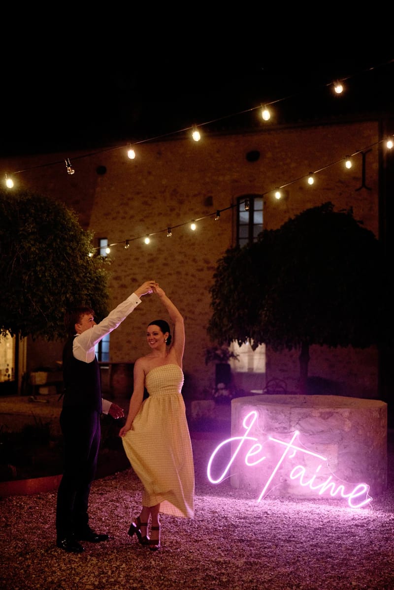 Groom spinning bride in yellow smocked dress under Edison lights with pink Je t'aime neon sign at night