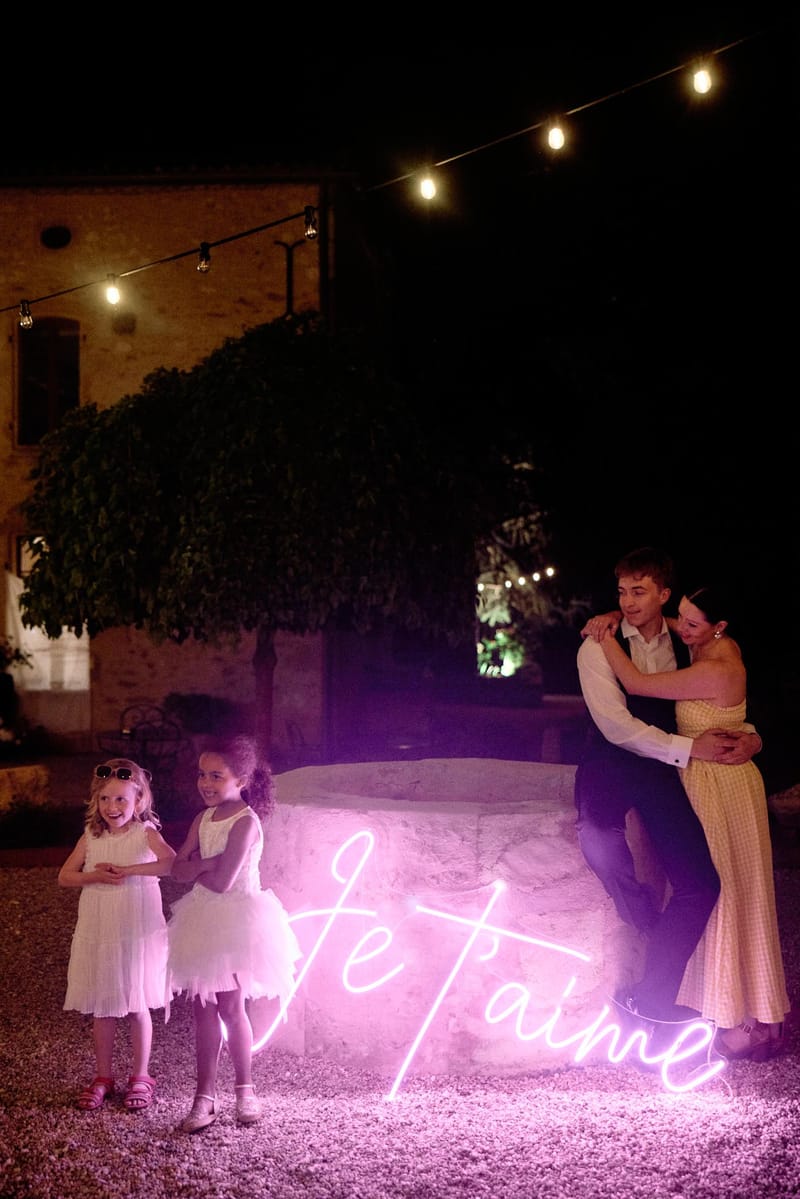Evening reception scene with pink neon Je t'aime sign on stone wall as guests gather in courtyard