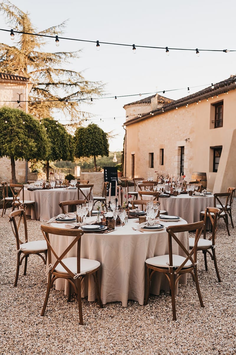 Round tables with burgundy blooms, dark tapers, and cross-back chairs under Edison lights in stone courtyard
